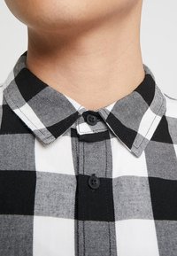 Camicia a quadri bianco e nero con colletto a punta, caratterizzata da una superficie liscia e un motivo a quadri contrastante. Due bottoni neri visibili.