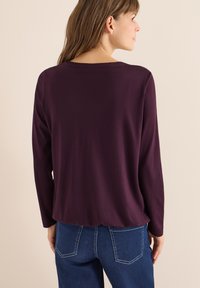 Langsleeve top in dieppaars stof met een relaxte pasvorm, voorzien van een brede halslijn en een gebogen zoom, gedragen met blauwe denim broek.