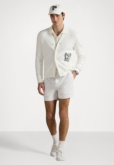Gilet blanc en tricot texturé, logo brodé vert, chemise blanche et short blanc, associé à une casquette blanche et des baskets.