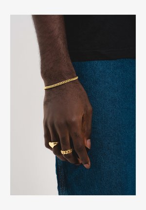 Tamsios odos ranka su auksine grandinėlės apyranke ir dviem auksiniais žiedais, vienas jų širdies formos, padėta šalia mėlynų džinsų.