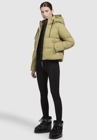 Jachetă puf în verde măsliniu, cu glugă, închidere cu fermoar și buzunare laterale. Asortată cu colanți negri și botine negre lucioase.