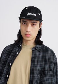 Black cap with "Service" embroidered in white, worn with a tan T-shirt and a black plaid shirt. Displayed on a plain background.