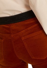 Pantalon couleur rouille avec une texture douce, doté d'une taille élastique noire avec logo, de poches arrière et de détails de couture subtils.