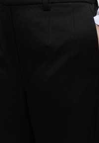 Gros plan d'une personne portant un pantalon noir sur mesure avec une poche latérale et une manche de chemise blanche visible à droite.
