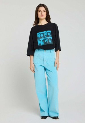 Jeune femme debout portant un top court noir avec un collage photo bleu et un pantalon large taille haute bleu clair avec des chaussures noires.