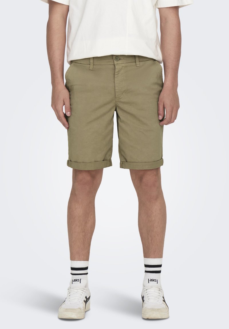 Shorts kaki avec une coupe classique, des ourlets roulés et un design à plat. Associés à des baskets blanches et des chaussettes à rayures noires.