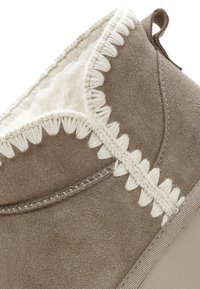 Bruine suède pantoffel met een witte schapenvoering, decoratieve gevlochten stiksel en een getextureerde zool. Beschikt over een achtertab voor gemakkelijk aan- en uittrekken.