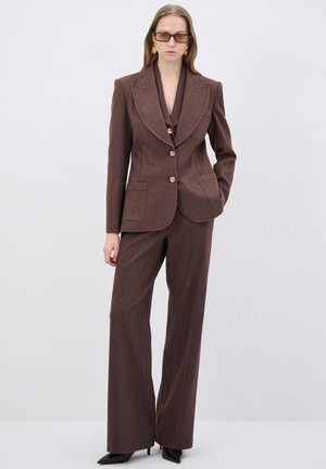 Bruine op maat gemaakte suit met een blazer met notch-lapel en bijpassende wijde broek. Kenmerkt zich door de gestructureerde stof en gouden knopen.