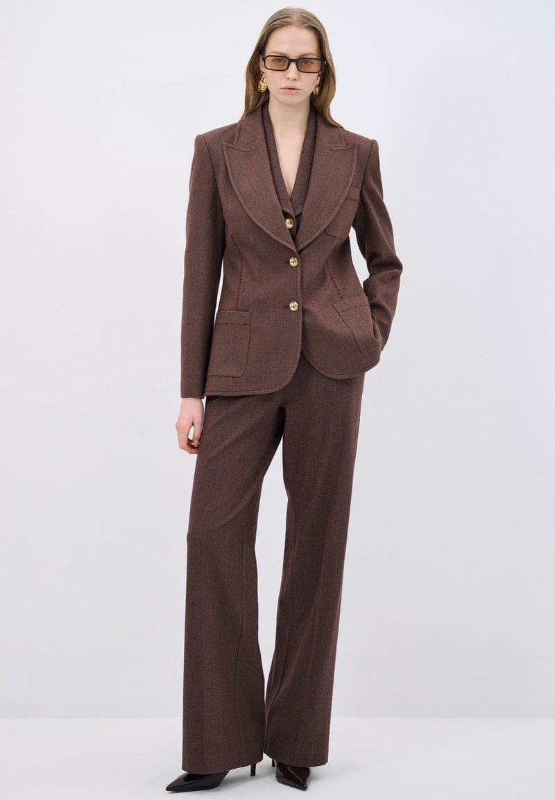 Bruine op maat gemaakte suit met een blazer met notch-lapel en bijpassende wijde broek. Kenmerkt zich door de gestructureerde stof en gouden knopen.