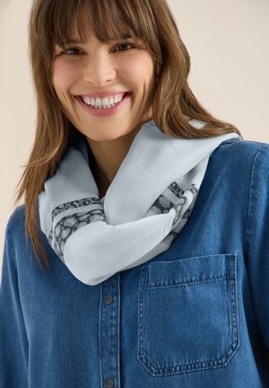 Giovane donna con capelli castani che sorride, indossando una sciarpa azzurra con motivi e una camicia di jeans blu con bottoni.