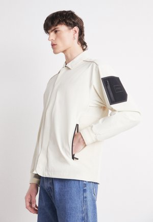Light beige jacket with a collared neckline, side pockets, and a black zippered pocket on the left arm. Smooth texture and minimal design.