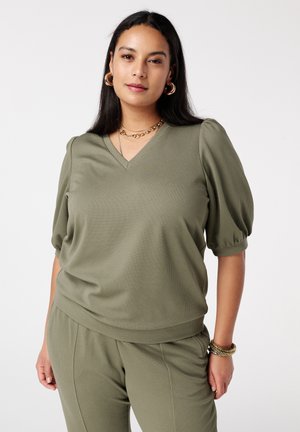 Femme portant un haut vert olive texturé à col en V avec manches bouffantes et pantalon assorti, accessoirisée avec des boucles d'oreilles créoles en or et des colliers superposés.