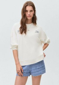 White sweatshirt with text, relaxed fit, and dropped shoulders paired with blue lace shorts featuring scalloped edges and textured layers.