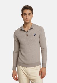 Ein junger Mann trägt einen beigen Pullover mit Viertelreißverschluss und kleinem gesticktem Logo, kombiniert mit hellen Hosen, und steht vor einem weißen Hintergrund.