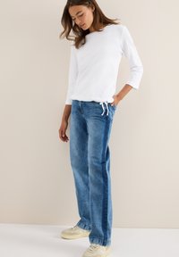 Haut à manches longues blanc associé à un jean en denim droit bleu clair avec des rayures latérales, porté avec des baskets beiges. Fond uni.