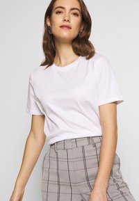Weißes T-Shirt mit Rundhalsausschnitt und kurzen Ärmeln, aus weicher Baumwolle. Getragen mit hellgrauen karierten Hosen mit blauen und braunen Linien.