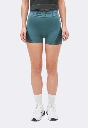 Pige iført turkis kompressionsshorts med høj talje, hvide ankelstrømper og hvide Nike-sneakers stående foran en ensfarvet baggrund.