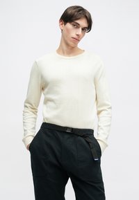 Ανοιχτόχρωμο πουλόβερ με στρογγυλή λαιμόκοψη, μακριά μανίκια και ραμμένα μανίκια; συνδυασμένο με σκούρους, φαρδύς παντελόνες που διαθέτουν ζώνη και τσέπες.