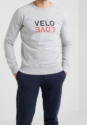 Man draagt een lichtgrijze sweatshirt met zwarte "VELO" en omgekeerde rode "LOVE" tekst, gecombineerd met donkerblauwe broek, handen in zakken.