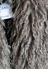 Struktureret, krøllet uldfrakke i nuancer af brun og grå. Etiketten synlig, der står "produceret i Italien." Blød, fluffy udseende med definerede vridninger.