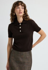 Žena s kudrnatými vlasy nosí krátkosrstý tmavě hnědý svetr s perlovými knoflíky a sukni z tweedové látky s vysokým pasem, která má multicolorové nitě.