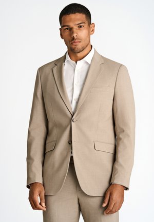 Man met beige colbert en bijpassende broek en een wit overhemd, staand tegen een effen witte achtergrond, gezicht naar voren gericht.