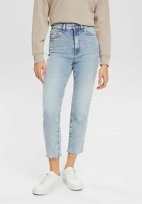 Jean taille haute bleu clair en denim, avec une coupe droite et un ourlet effiloché. Assorti à des baskets blanches et un pull beige.