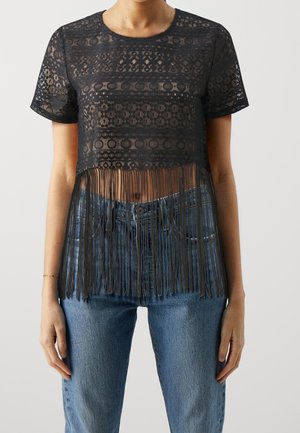 Czarny przezroczysty krótki top z koronki z krótkimi rękawami i długim frędzlowanym dołem, noszony z niebieskimi dżinsami, odsłaniający talię i pępek.