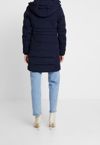 Giacca imbottita blu navy con cappuccio, texture trapuntata, lunghezza lunga e polsini elasticizzati, abbinata a jeans azzurri chiari e stivali con tacco beige.
