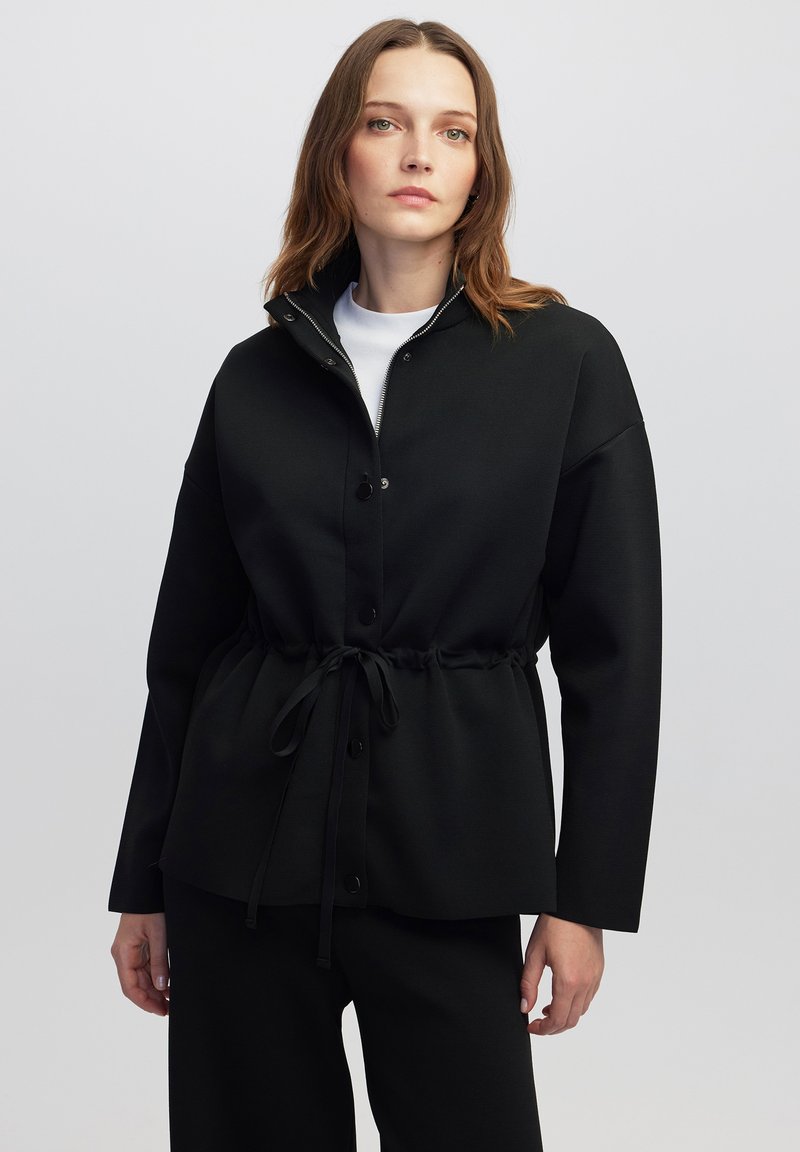 Zwarte, loszittende jas met een kraag, knoopsluiting aan de voorkant en een verstelbare trekkoord in de taille. Glad textiel, lange mouwen en een minimalistisch ontwerp.