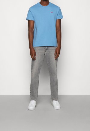 Mężczyzna stojący, ubrany w jasnoniebieską koszulkę z krótkim rękawem, szare dżinsy i białe tenisówki na jednolitym tle.