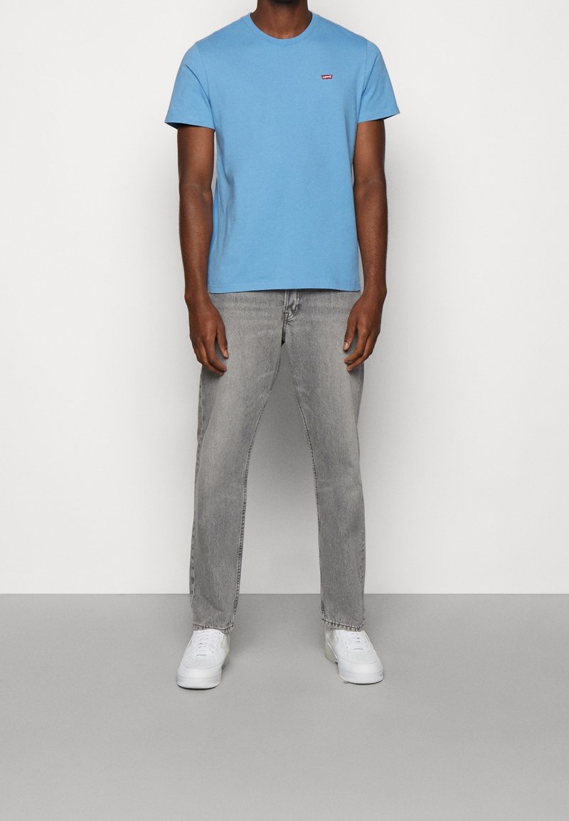 Homme debout portant un T-shirt à manches courtes bleu clair, un jean gris et des baskets blanches, sur un fond uni.