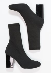 Paire de bottines chaussettes noires en tissu maille extensible avec talons blocs brillants présentées sur un fond blanc.