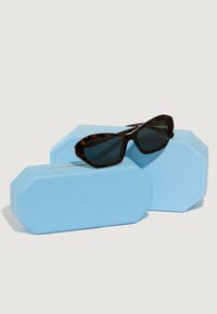 Occhiali da sole tartarugati con lenti scure, poggiati su due custodie ottagonali di colore blu chiaro, con superficie texturizzata e logo.