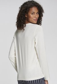 Langärmliges Oberteil mit Rundhalsausschnitt aus einem hellen Creme-Stoff mit glatter Textur. Hat eine lockere Passform und seitliche Schlitze am Saum.