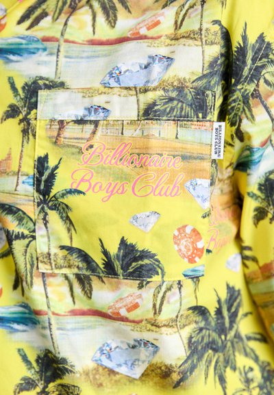 Cămașă cu mânecă scurtă din țesătură galbenă, cu un imprimeu tropical cu palmieri, apă și diamante; logo-ul "Billionaire Boys Club" în roz.