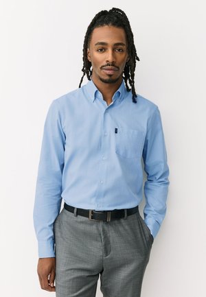 Mand med dreadlocks iført en lyseblå skjorte med knapper, stoppet ned i grå bukser, står foran en hvid baggrund.
