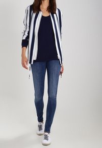 Jacka med dragkedja i blå och vita vertikala ränder, kombinerad med en marinblå topp och blå jeans, bärs med vita sneakers.