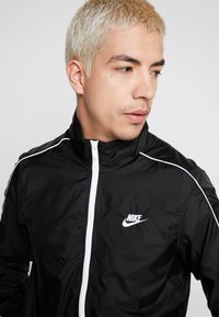 Jauns vīrietis ar platīna blondiem matiem, ģērbies melnā Nike jakā ar baltu apdari un rāvējspiedi, skatās pa labi uz vienkārša fona.