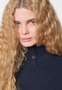 Junge Frau mit langen, welligen blonden Haaren, die einen marineblauen Strickpullover mit hohem Kragen und drei dunklen Knöpfen trägt.