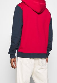 Hombre vistiendo una sudadera roja con mangas y cintura en azul marino, combinada con pantalones en color blanco roto, visto desde atrás.