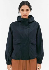 Svart vattentät jacka med huva, elastiska ärmslut och två framfickor. Slät tygkvalitet med avslappnad passform och minimalistisk design.