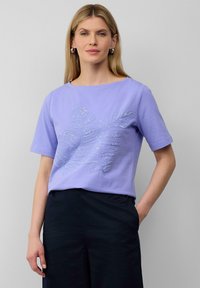 Lichtpaarse t-shirt met korte mouwen, voorzien van een decoratief zilverkleurig bladpatroon in het midden. Gekoeld met donkere broeken.