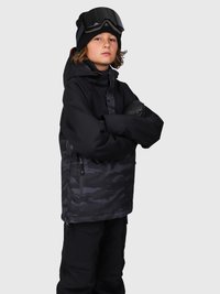Niño vestido con una chaqueta de esquí negra con patrón de camuflaje, guantes negros, pantalones negros, un gorro negro y gafas de esquí en la cabeza, de pie con los brazos cruzados.