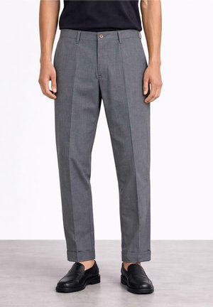 Mężczyzna w szarych spodniach wizytowych z mankietami, czarnych mokasynach i w czarnej koszuli włożonej do środka, stojący na szarej podłodze na białym tle.