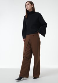 Schwarzer Rollkragenpullover mit weiten Ärmeln, kombiniert mit hochgeschnittenen braunen Hosen mit Falten und Seitentaschen. Schwarze spitze Absätze.