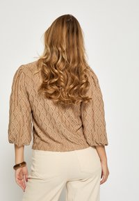 Brun tekstureret bluse med diamantmønster, trekvart puffede ærmer og en afslappet pasform, stylet med cremefarvede bukser.