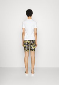 Vit t-shirt med kort ärm som är kombinerad med svart och guld-mönstrade shorts med utsmyckade mönster, bärs tillsammans med vita slip-on-skor.