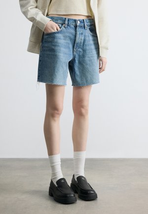 Személy világoskék farmer rövidnadrágot visel, rojtos szegéllyel, fehér zoknit, fekete platform loafer cipőt, bézs kivágott felsőt és bézs cipzáros kapucnis pulóvert.