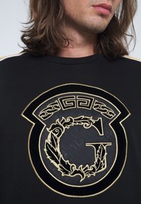 Homme aux cheveux mi-longs portant une chemise noire avec un grand emblème décoratif en or et noir en forme de lettre "G" sur la poitrine.